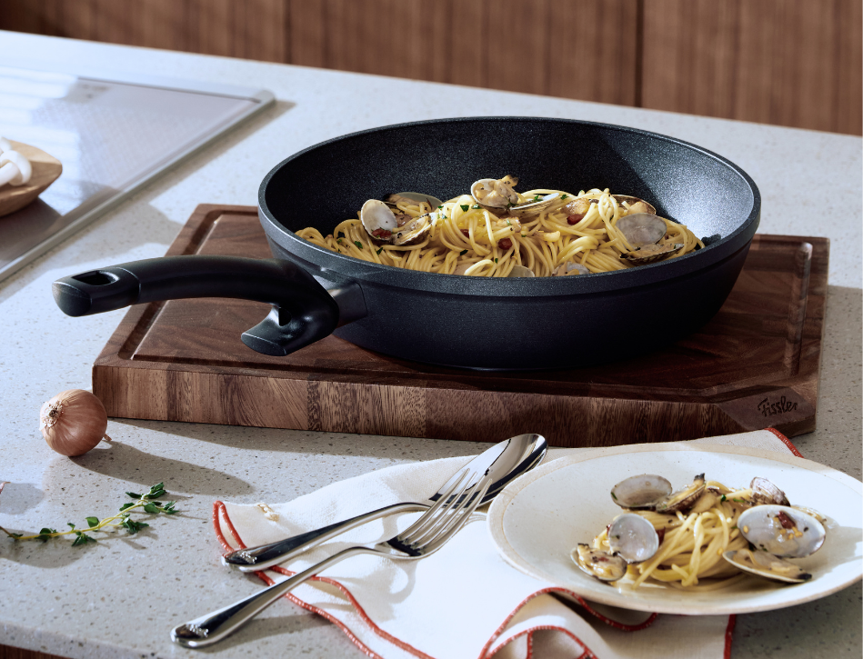 Auf einer schwarzen Fissler-Pfanne, die fachmännisch auf Langlebigkeit und Leistungsfähigkeit gefertigt wurde, sind Spaghetti mit Venusmuscheln auf einem Holzschneidebrett angerichtet. Daneben steht auf der Küchenarbeitsplatte eine Portion Pasta auf einem Teller, flankiert von hochwertigem Besteck, frischen Zwiebeln und duftenden Kräutern – ein Beispiel für zeitloses deutsches Design und kulinarische Innovation.