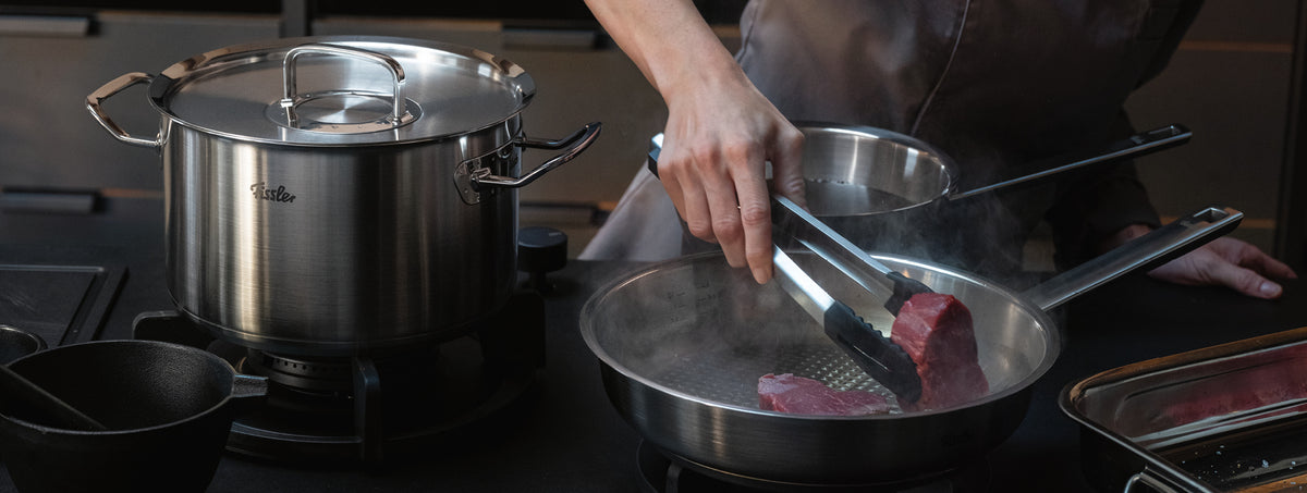 Ein Koch legt Steaks in eine Fissler-Edelstahlpfanne, die neben einem passenden Topf auf dem Herd steht. Präzision, Dampf und Innovation glänzen.