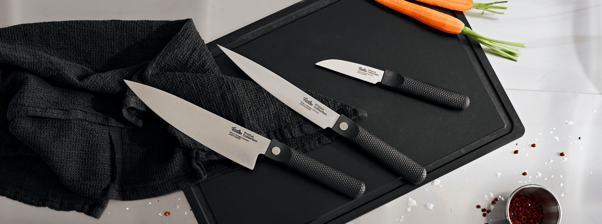 Fissler Präzisionsküchenmesser auf einem schwarzen Brett, daneben Karotten, Handtuch, Salz, Pfefferkörner und eine kleine Tasse mit rotem Gewürz.