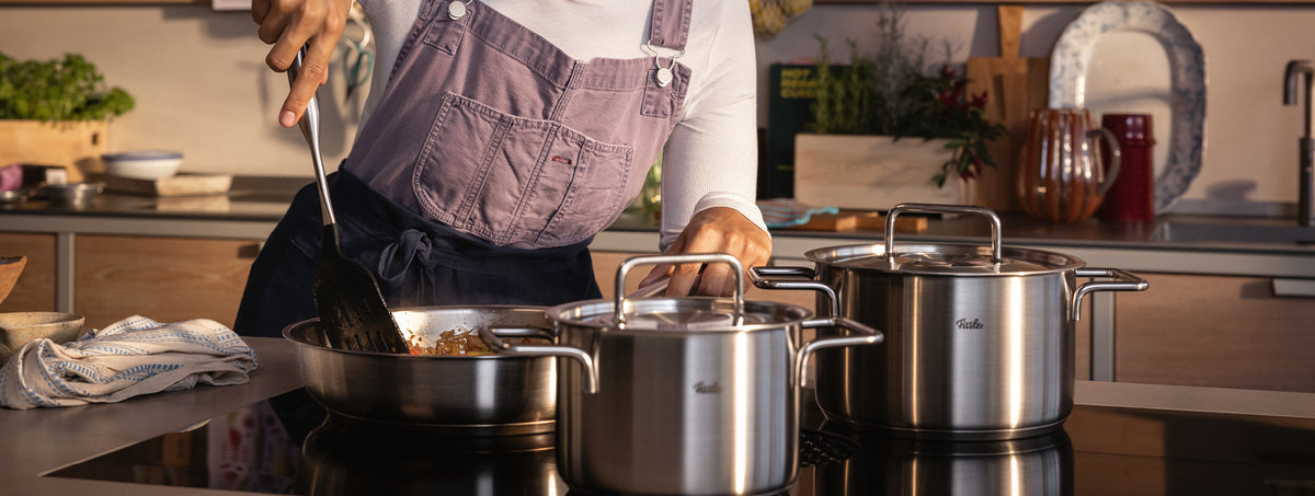Eine Köchin in rosa Schürze kocht mit Fissler-Edelstahltöpfen auf dem Herd; Grünzeug und hochwertiges Kochgeschirr schmücken die Küche.