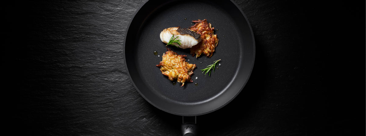 Eine Fissler Premium-Bratpfanne mit gekochtem Fisch, frischen Kräutern und goldenen Kartoffelrösti, die deutsche Qualitätsarbeit zeigt.