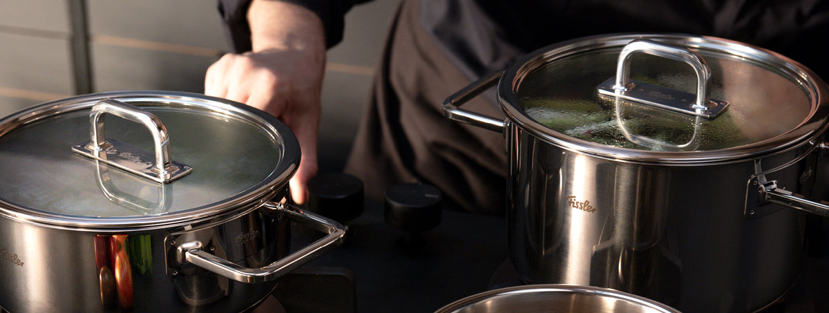 Ein Koch am Herd kümmert sich um zwei Fissler-Edelstahltöpfe mit Glasdeckeln, die in Deutschland präzisionsgefertigt wurden, während im Inneren Dampf aufsteigt.