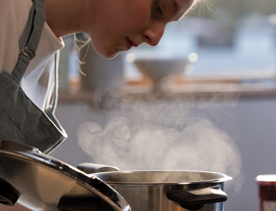 A female chef in an apron lifts the lid of a Fissler stainless steel pot, enjoying the aromatic steam in a bright, modern kitchen.