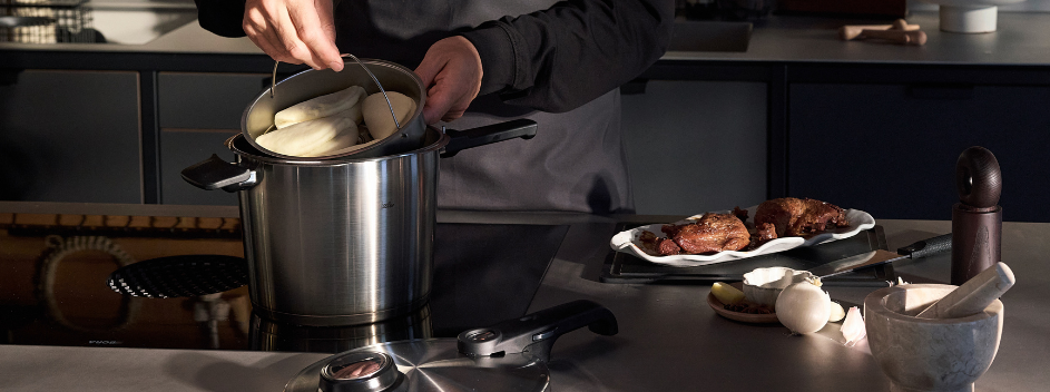 Eine Person legt geschältes weißes Gemüse in einen Fissler-Edelstahlkorb, den sie in einen hochwertigen Fissler-Topf auf dem Herd stellt, und veranschaulicht damit die Präzision und Innovation von Kochgeschirr aus deutscher Fertigung. Neben dem Topf liegen auf der Arbeitsplatte gegartes Fleisch, frische Zwiebeln und eine Pfeffermühle – ein Zeichen für Fisslers Engagement für Qualität und zeitloses Design für jeden Moment in der Küche.