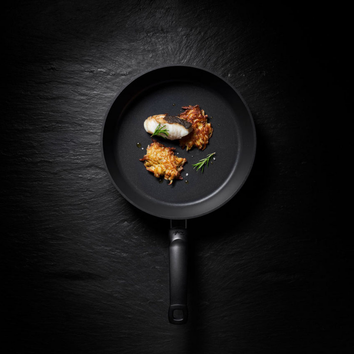 A Fissler frying pan on a dark surface, elegantly serving fish, potato pancakes, and rosemary – a testament to German quality and design.