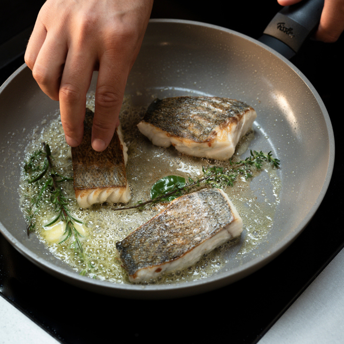 Eine Hand legt behutsam ein Fischfilet mit knuspriger Haut in eine hochwertige Fissler-Pfanne, wo sprudelnde Butter und frische Kräuter zusammen mit zwei weiteren goldbraunen Filets auf dem Herd für Geschmack sorgen – ein Beispiel für präzises Kochen in zeitlosem, in deutscher Ingenieurskunst gefertigtem Kochgeschirr.