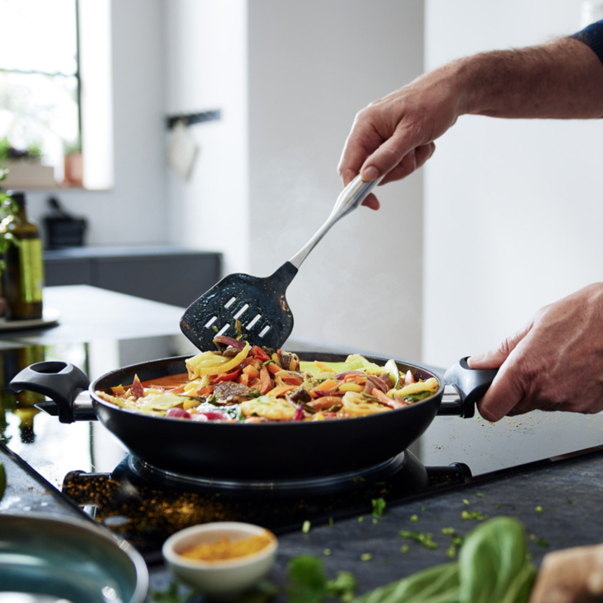 In einer modernen Küche brät eine Person mit einem Pfannenwender Gemüse und Nudeln in einer eleganten schwarzen Fissler-Pfanne auf dem Herd an und unterstreicht damit die deutsche Präzision und das zeitlose Design der Marke.