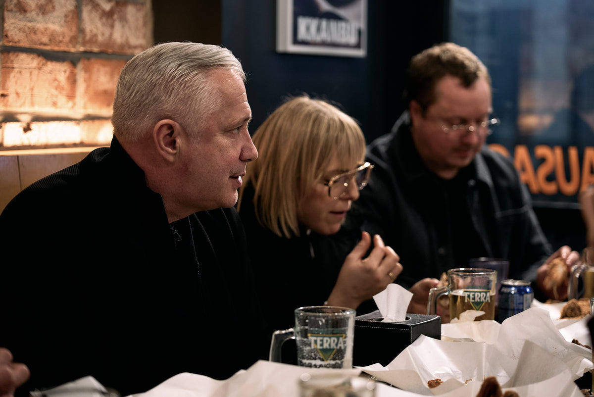 Drei Personen genießen gemeinsam eine Mahlzeit in einem Restaurant und sitzen um einen Tisch herum, auf dem Essen und Getränke stehen. Zwei von ihnen tragen eine Brille, und alle sind in dunkler Kleidung gekleidet, die einen zeitlosen Stil widerspiegelt. Im Hintergrund sind eine Backsteinmauer und leicht unscharf wirkende Schilder zu sehen. Hochwertiges Kochgeschirr, wie beispielsweise Edelstahltöpfe oder -pfannen von Fissler, könnte für die Zubereitung der Gerichte, die sie genießen, verwendet worden sein