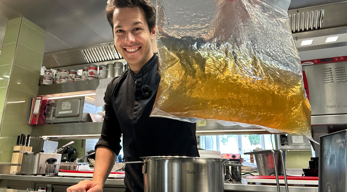 Ein Koch in einer modernen Küche lächelt, während er eine große, durchsichtige Plastiktüte mit gelblicher Flüssigkeit über einen hochwertigen Fissler-Edelstahltopf hält und damit die präzise Technik und das zeitlose Design unseres hochwertigen Kochgeschirrs „Made in Germany“ präsentiert.