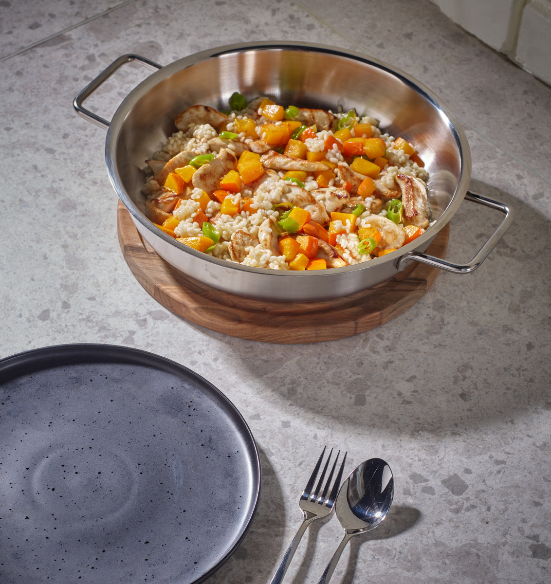 Fissler Pure Servierpfanne, Made in Germany aus recyceltem Edelstahl, gefüllt mit Reis, Gemüse, Huhn; Teller und Besteck daneben.