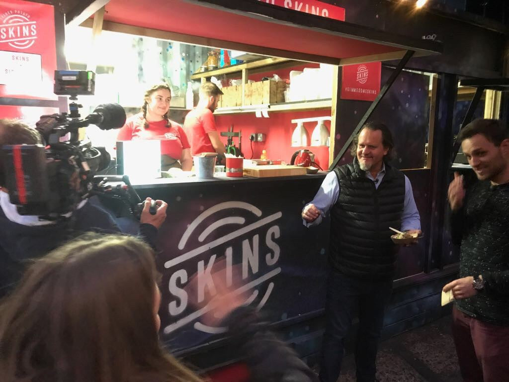 Ein Filmteam fängt die lebhafte Atmosphäre am Abend bei einem Food-Truck namens SKINS im Freien ein, wo zwei Mitarbeiter in roten Hemden köstliche Gerichte durch das Fenster servieren. Draußen unterhalten sich zwei Männer und lächeln einander zu – ein Ausdruck der Freude an guter Gesellschaft und meisterhaft zubereitetem Essen. Diese lebendige Szene spiegelt Fisslers Engagement für kulinarische Exzellenz wider – verwurzelt in der deutschen Tradition seit 1845 –, wobei hochwertiges Kochgeschirr aus