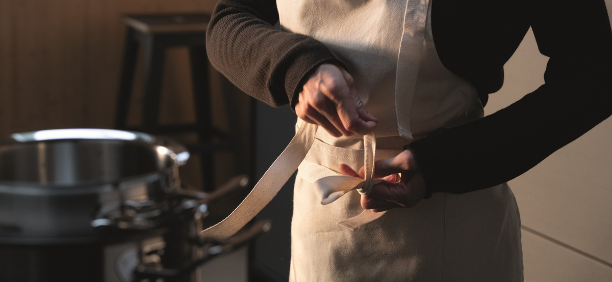 A person ties a beige apron in a kitchen, with a high-quality Fissler stainless steel pot and a stool in the background.