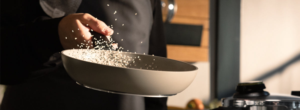 Eine Person schüttelt gekonnt weiße Körner – vermutlich Reis – in einer beigen Fissler-Pfanne über dem Herd hin und her und unterstreicht damit die präzise Technik der Pfanne, da einige Körner mitten in der Luft aufgefangen werden. Das zeitlose Design und die hochwertige Verarbeitung der Fissler-Kochgeschirre machen das tägliche Kochen zu einer Kunstform.