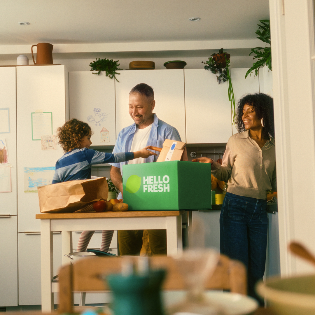A family of three gathers in a bright kitchen, smiling as they unpack fresh ingredients together beside a collection of premium Fissler stainless steel cookware displayed on the counter. Drawings are visible on the white cabinets in the background, adding warmth to the timeless space defined by precision-engineered pots and pans "Made in Germany.