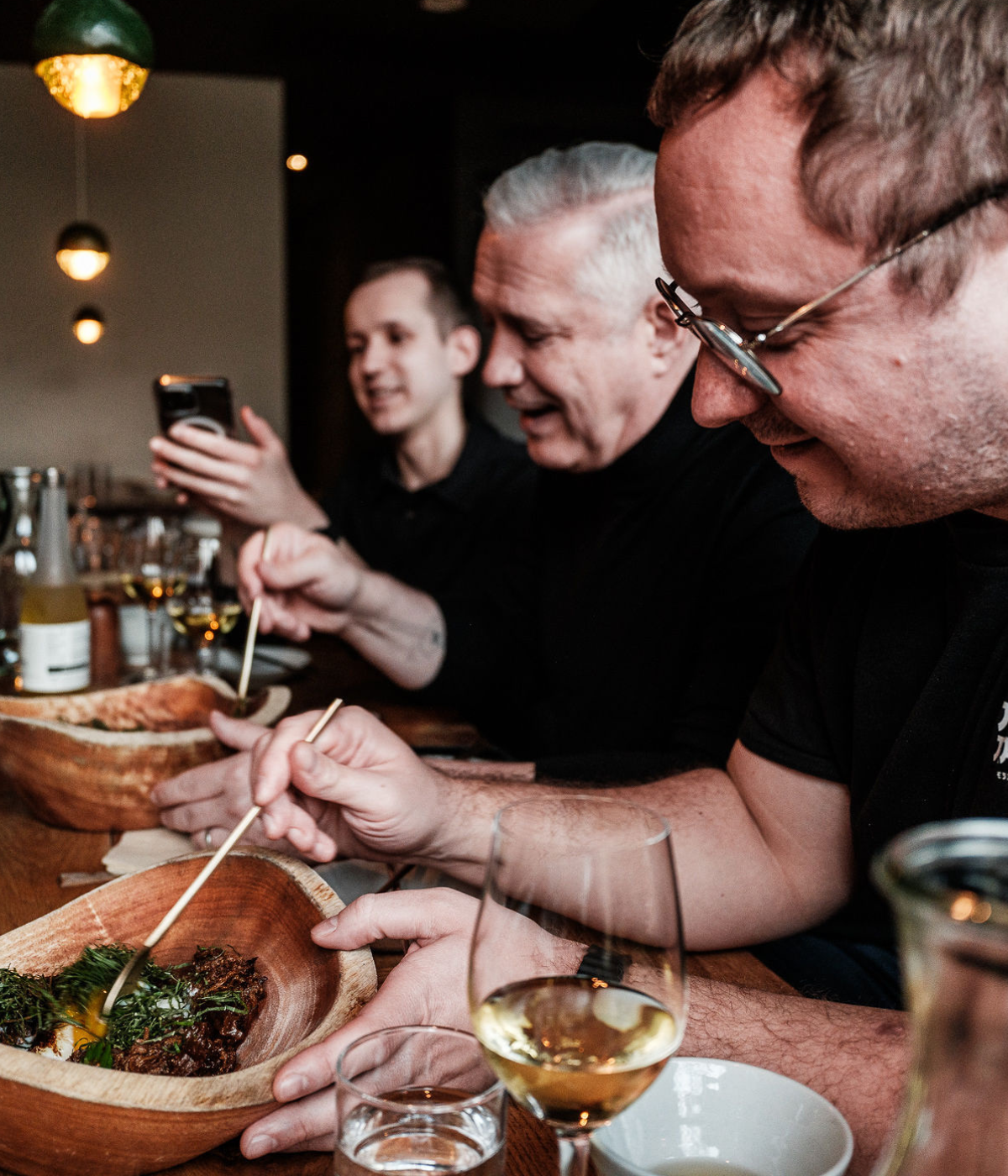 Three people enjoy a meal together at a table, sharing smiles and laughter. Freshly prepared dishes are served in wooden bowls, with drinks beside them. High-quality Fissler stainless steel cookware and elegant kitchen accessories complement the scene, reflecting German craftsmanship and timeless design. One person holds a phone while another eats with chopsticks, illustrating modern dining alongside precision-engineered Fissler products.