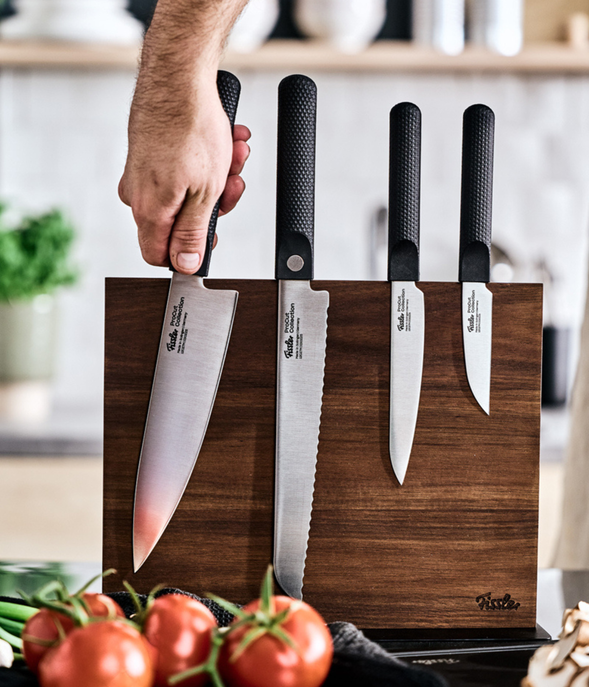 A hand selects a precision-engineered chef’s knife from a Fissler wooden block, showcasing four versatile Fissler knives. In the foreground, fresh tomatoes and aromatic herbs highlight the timeless design and German quality integral to Fissler’s innovative kitchen accessories.