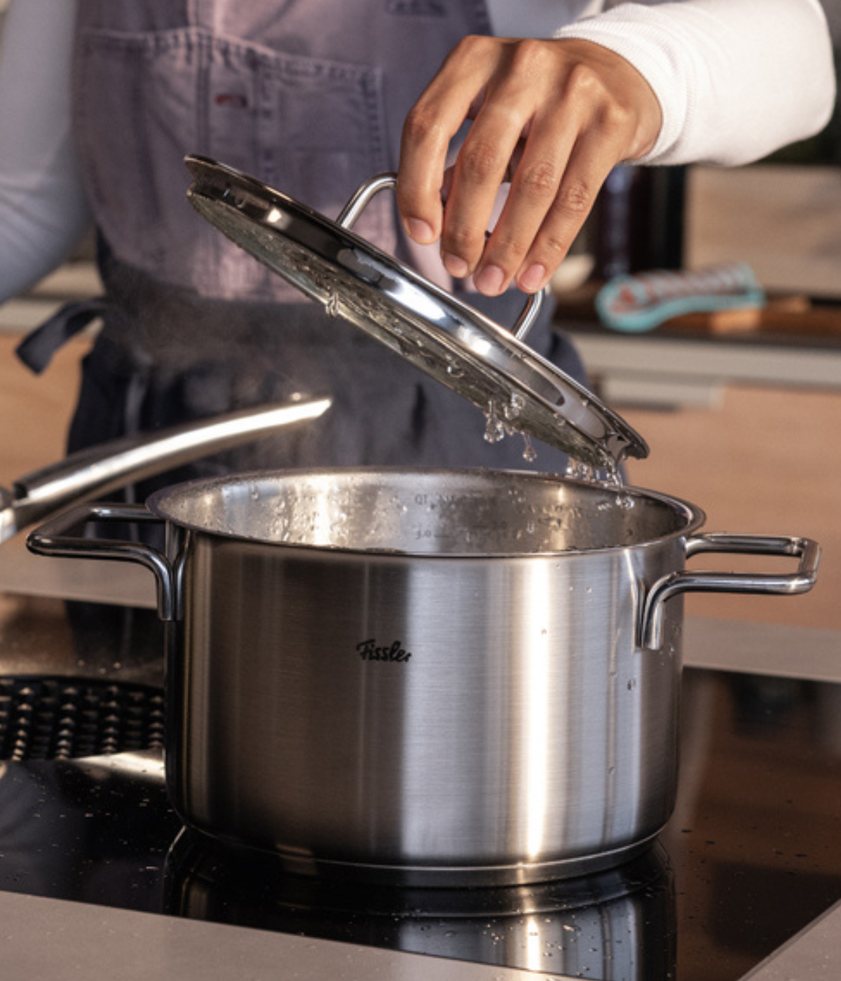 A person in a grey apron lifts the glass lid from a steaming Fissler stainless steel pot on a sleek black stovetop, showcasing German-made precision and innovation as condensation drips from the expertly engineered lid.