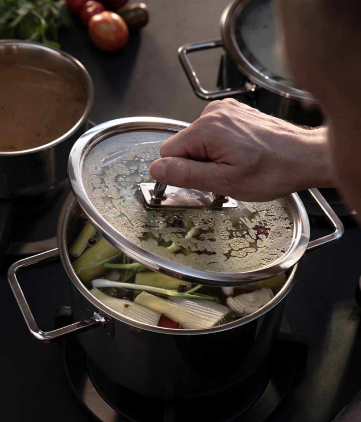 A close-up, high-angle shot highlights a hand lifting the premium glass lid from a Fissler stainless steel pot, expertly engineered for precise cooking. The pot is filled with vibrant vegetables and savory broth on a modern stovetop. Next to it sits another gleaming Fissler pot, while fresh tomatoes and herbs provide a colorful accent in the softly blurred background—reflecting our heritage of innovative German craftsmanship since 1845.