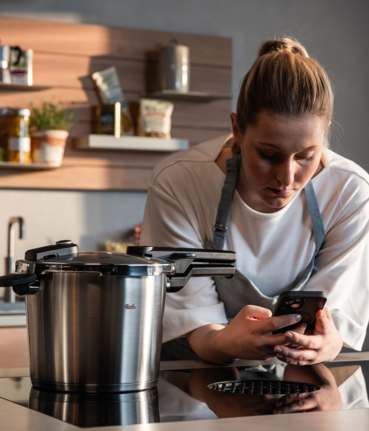 A woman in an apron leans on a kitchen counter, looking at her phone beside a premium Fissler stainless steel pressure cooker on the stove. Shelves with neatly organized jars and ingredients highlight the well-appointed kitchen, reflecting Fissler’s tradition of German engineering, timeless design, and durable cookware.
