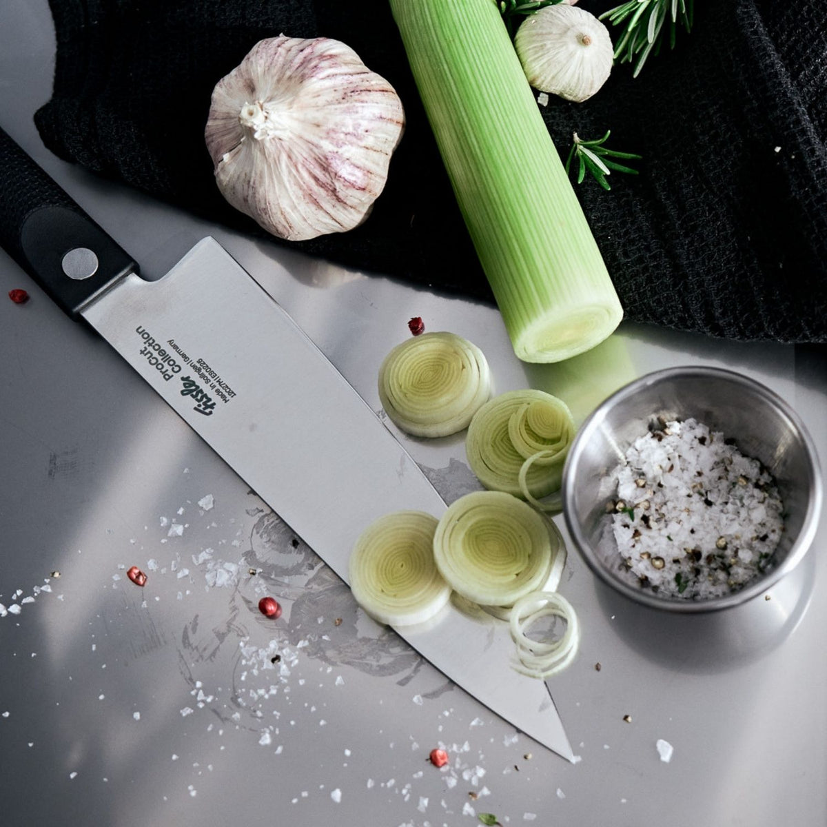 A precise Fissler stainless steel chef's knife with fresh leeks, garlic, rosemary, and a steel cup of coarse salt and pepper.