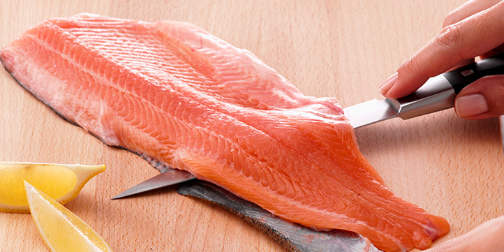 Eine Person filetiert frischen Lachs auf einem Holzbrett mit Zitronenscheiben, wobei ein Fissler-Premiummesser für Präzision und Qualität verwendet wird.