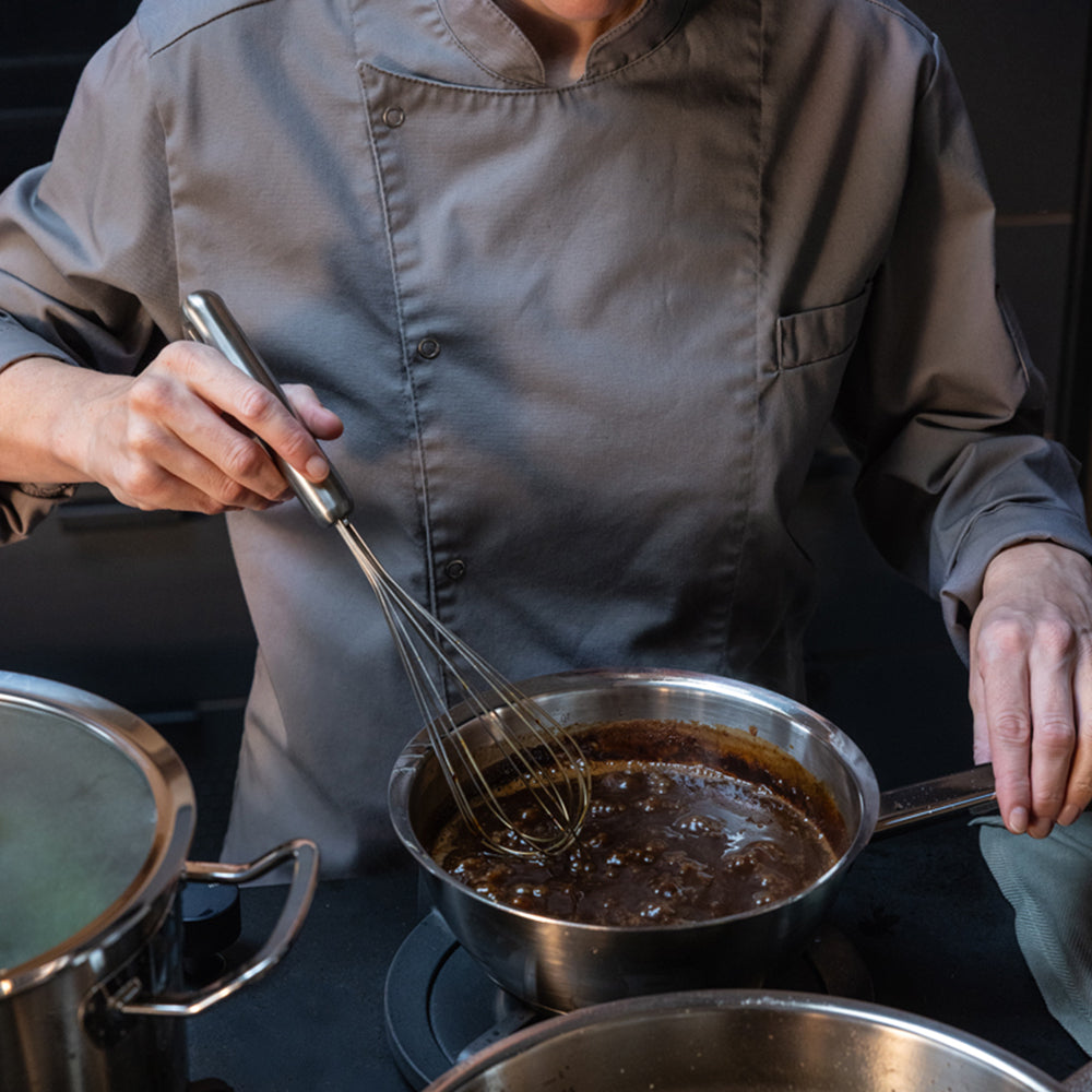Ein Koch in grauer Uniform rührt gekonnt mit einem Schneebesen eine cremige Soße in einem hochwertigen Fissler-Edelstahltopf auf dem Herd um und demonstriert damit präzise Handwerkskunst und zeitloses deutsches Design. Daneben steht ein weiterer Fissler-Topf, der das Bekenntnis der Marke zu Innovation und Langlebigkeit in Profiküchen widerspiegelt.