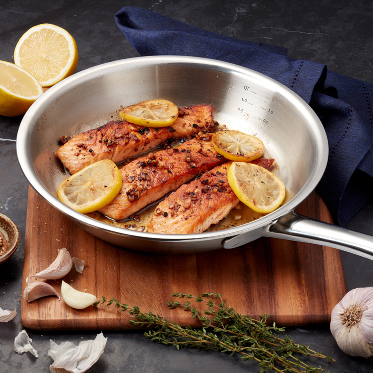 Meisterhaft zubereitete Lachsfilets mit Zitronenscheiben und aromatischen Gewürzen werden wunderschön in der Fissler M5 Pro-Ply® 3-tlg. Set mit Metalldeckel aus Edelstahl präsentiert und unterstreichen damit unsere renommierte deutsche Ingenieurskunst und unser zeitloses Design. Die Pfanne ruht auf einem Holzbrett, umgeben von frischen Knoblauchzehen, Thymianzweigen, einer halbierten Zitrone und einer eleganten dunkelblauen Serviette – ein Spiegelbild der Vielseitigkeit und Qualität, die