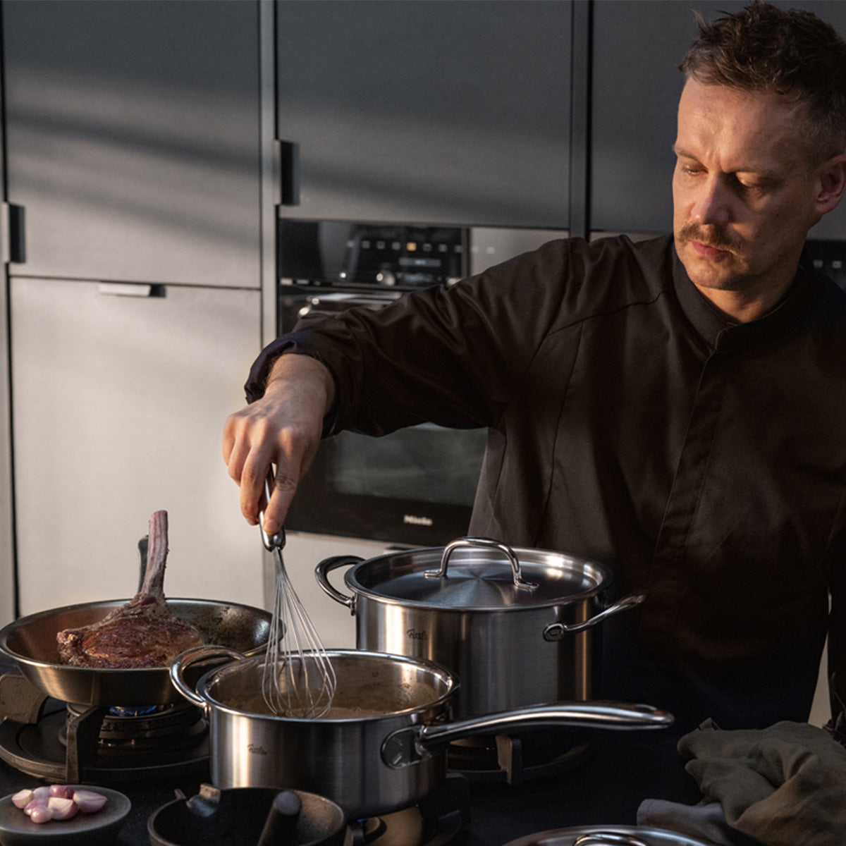 Ein Mann in einem dunklen Hemd kocht in einer modernen Küche und verrührt gekonnt eine Soße in einem Fissler M5 Pro-Ply®-Topf mit Metalldeckel, der Teil des hochwertigen 4-teiligen Sets ist, das für seine präzise Technik und zeitlose „Made in Germany“-Qualität bekannt ist. Neben ihm brutzelt ein Steak in einer weiteren Fissler-Pfanne aus derselben Kollektion. Dieses Kochgeschirr ist aus recyceltem Edelstahl gefertigt und für Induktionskochfelder geeignet – es überzeugt durch herausragende Langlebigkeit,