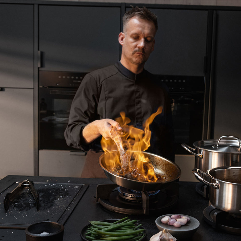 Ein Koch in schwarzer Kochuniform flambiert gekonnt Speisen in einer Fissler M5 Pro-Ply®-Pfanne aus dem hochwertigen 4-teiligen Set mit Metalldeckeln, das aus recyceltem Edelstahl gefertigt und ideal für Induktionskochfelder geeignet ist. Sichtbare Flammen steigen empor, während der Koch am Herd deutsche Präzision und Innovation demonstriert, umgeben von frischem Gemüse, einem weiteren Topf aus der Fissler-Kollektion und ausgewählten Kochzutaten auf der Arbeitsplatte – ein Beispiel für Fisslers Engagement