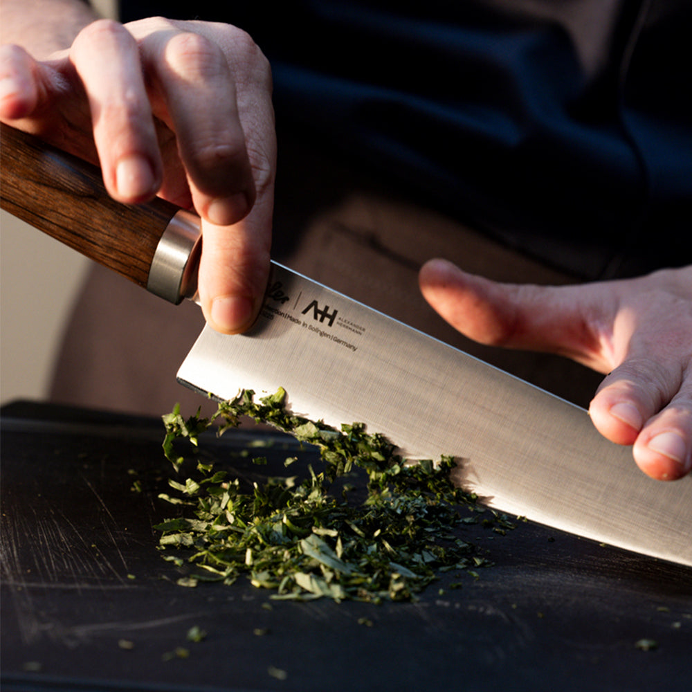 Eine Person schneidet gekonnt frische Kräuter auf einem eleganten schwarzen Schneidebrett mit einem hochwertigen, präzisionsgefertigten Fissler-Küchenmesser. Ihre Finger führen die Klinge sicher und souverän, was in jedem Detail die Tradition der Innovation und die zeitlose deutsche Qualität von Fissler widerspiegelt.
