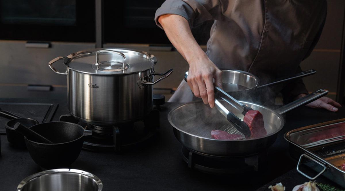 Ein Koch in einer Kochjacke brät gekonnt ein Stück Fleisch mit einer Zange in einer Fissler-Edelstahlpfanne an. Daneben steht ein großer Fissler-Topf auf dem Herd, umgeben von hochwertigen Küchenutensilien auf einer eleganten, dunklen Arbeitsplatte – ein Beispiel für die präzise Technik und das zeitlose Design der in Deutschland hergestellten Fissler-Kochgeschirre.