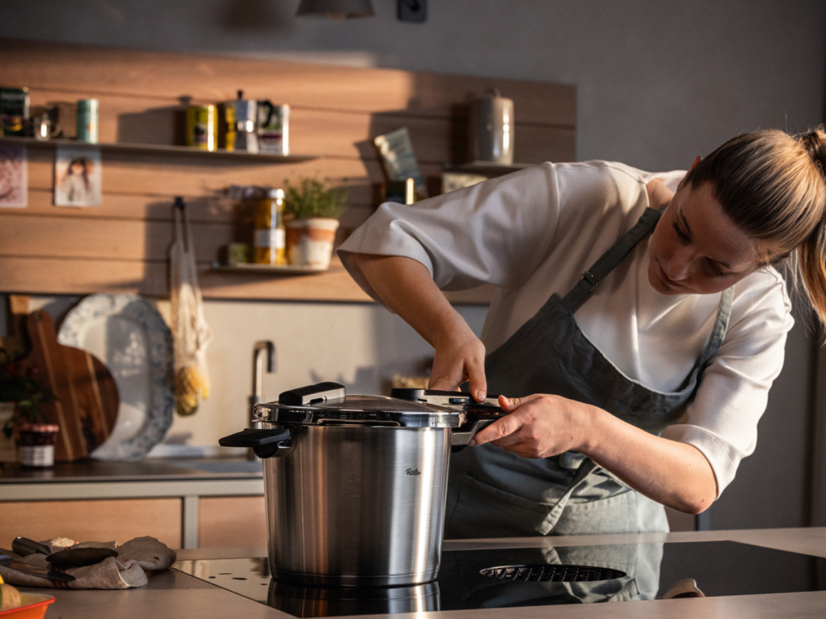 Eine Frau in einer Schürze steht in einer modernen Küche und schließt aufmerksam den Deckel eines Fissler-Edelstahl-Schnellkochtopfs auf dem Herd. Im Hintergrund sind Regale mit Gläsern, Pflanzen und Küchenaccessoires zu sehen, die die präzise Technik und das zeitlose Design der Fissler-Kochgeschirre unterstreichen.