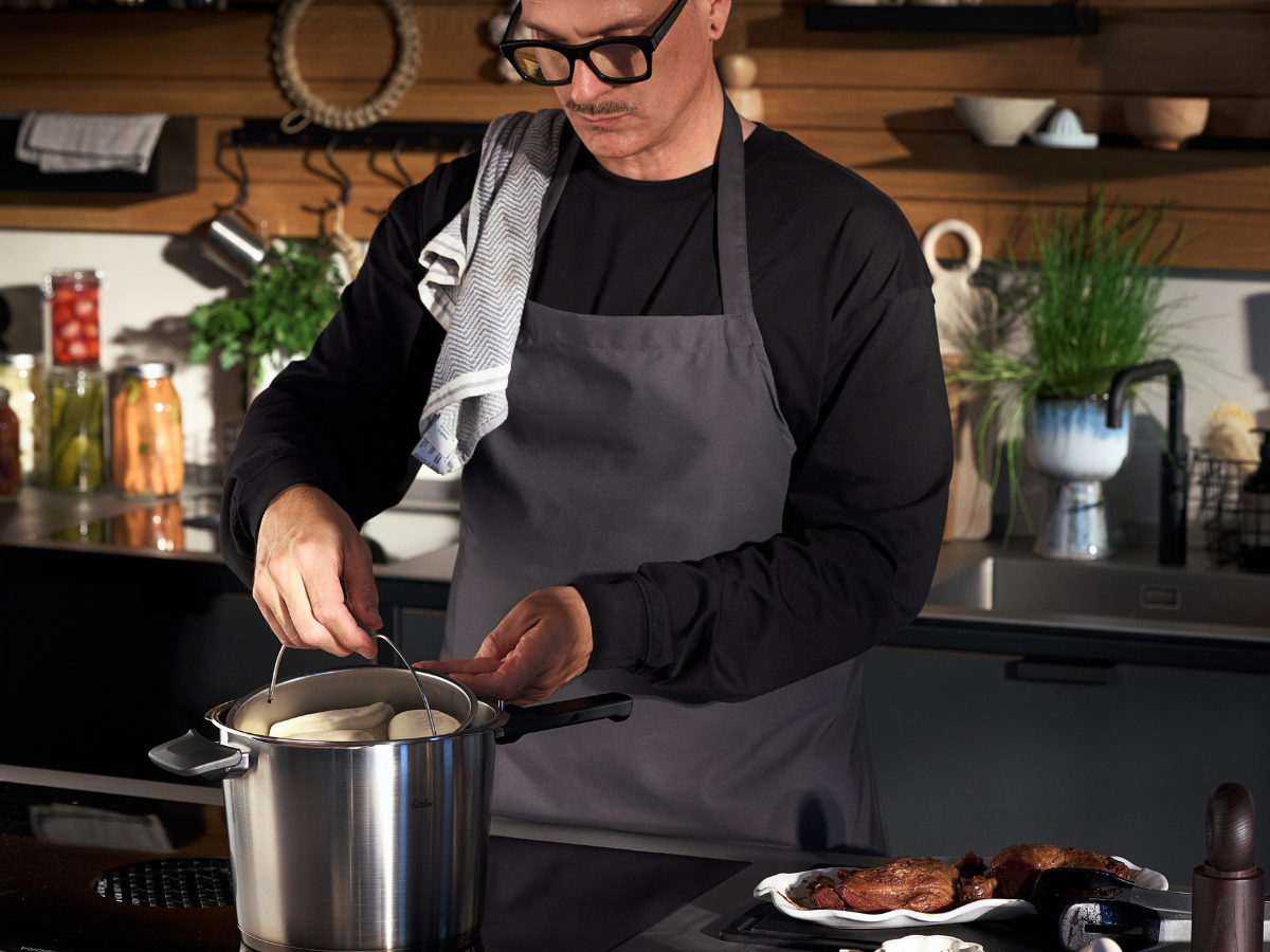 Eine Person mit Brille und grauer Schürze hebt den Deckel eines hochwertigen Fissler-Edelstahltopfs auf einem Herd in einer eleganten, modernen Küche an. Auf der Arbeitsplatte liegt das gegarte Fleisch elegant auf einem Teller neben dem Kochgeschirr und zeugt von dauerhafter Qualität und präziser Technik.