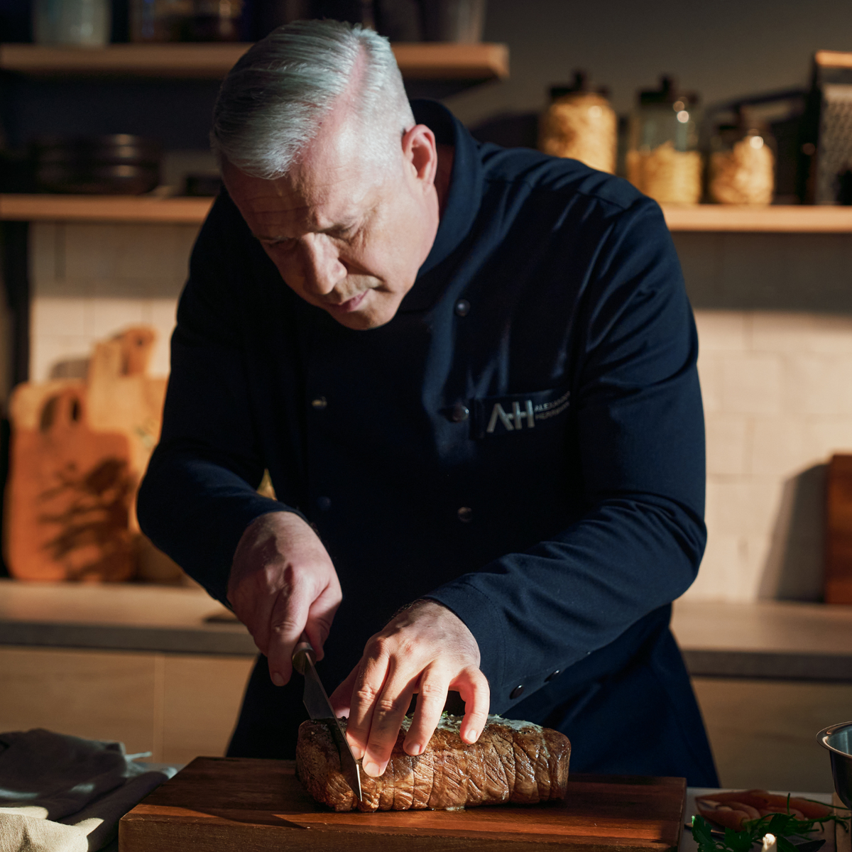 A chef with gray hair cuts a roast on a wooden board with a Fissler precision knife, with cookware and pots in the background.