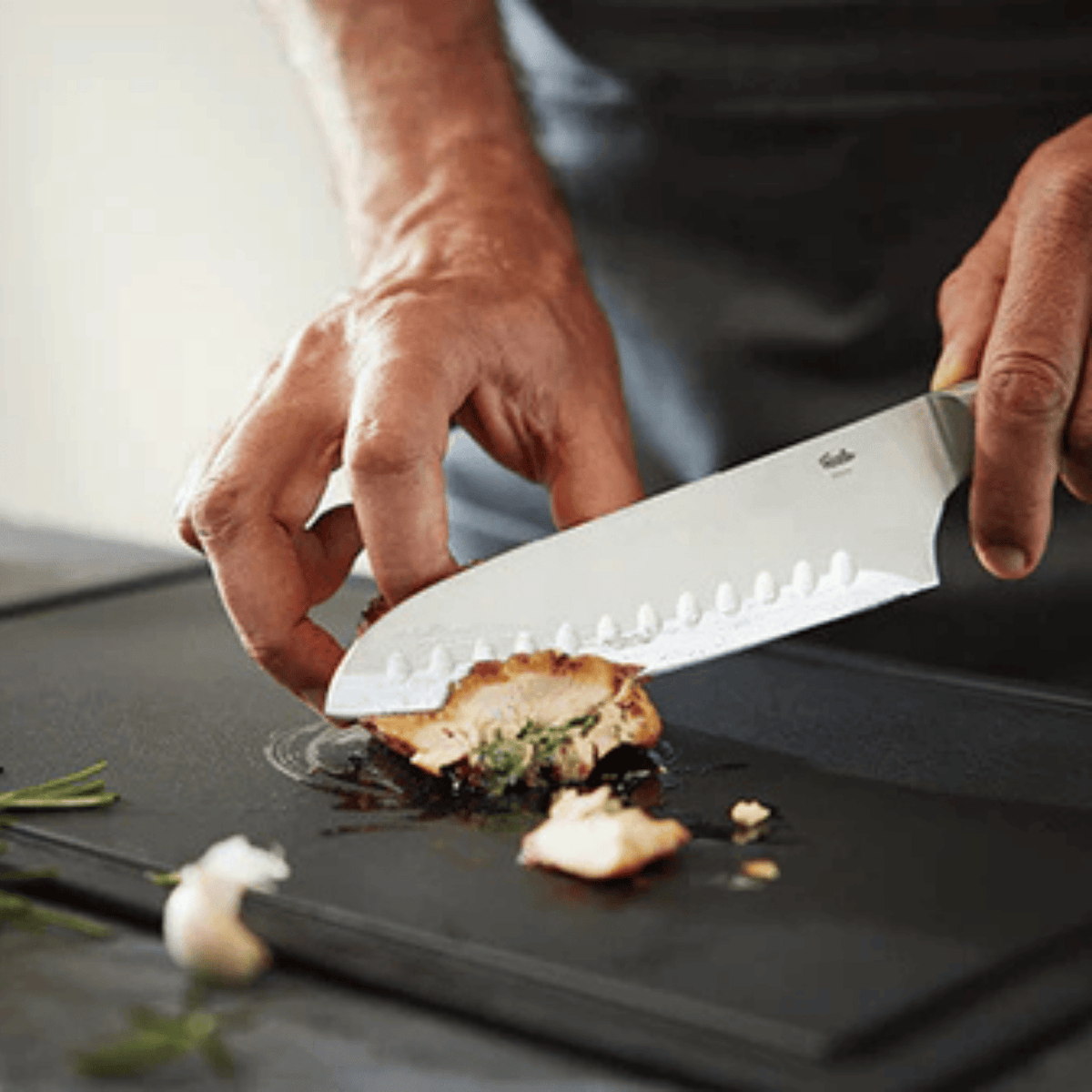 Eine Person schneidet gefüllte Hähnchenbrust mit einem präzisen Fissler-Kochmesser auf einem Schneidebrett auf, Kräuter und Knoblauch liegen daneben.