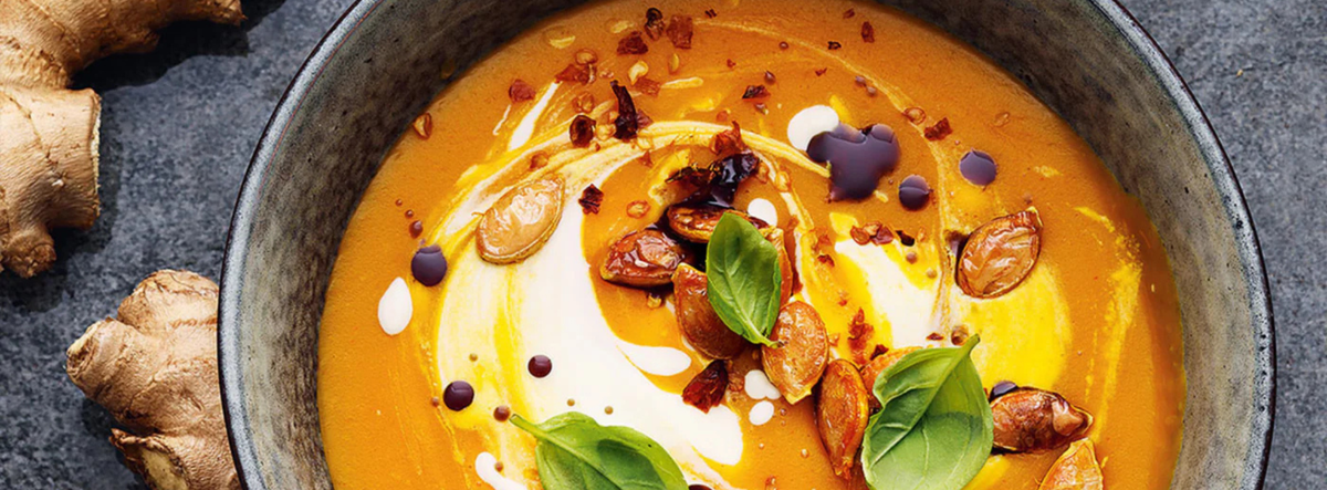 A bowl of rich, creamy orange soup elegantly presented with basil leaves, seeds, delicate swirls of cream, and a touch of oil, accompanied by fresh ginger pieces on a dark surface. Prepared and served using Fissler’s precision-engineered cookware for optimal flavor and timeless style.