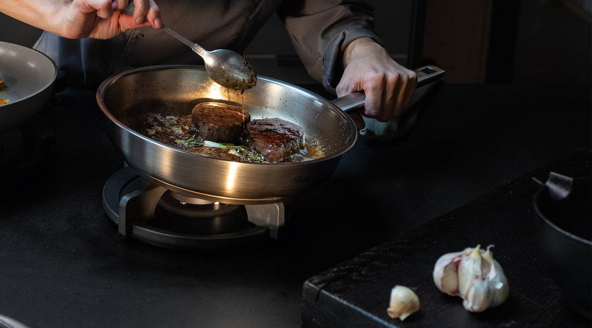 Eine Person brät gekonnt zwei Steaks in einer hochwertigen Fissler-Edelstahlpfanne an, gibt Butter über das Fleisch, während frische Kräuter und Knoblauch in der Pfanne brutzeln; ein ganzer Knoblauchknollen liegt auf der Arbeitsplatte und unterstreicht die Präzision und Qualität der Fissler-Kochgeschirre „Made in Germany“.