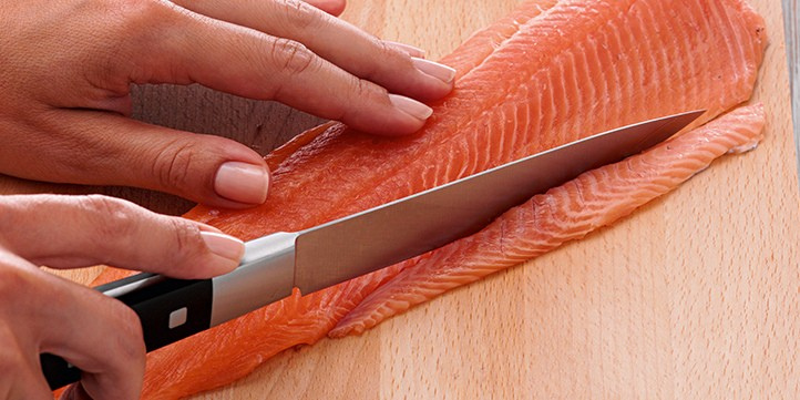 Mit einem Fissler-Präzisionsmesser schneidet eine Person fachmännisch frischen Lachs auf einem Holzbrett in Scheiben - zeitlose deutsche Qualität in Aktion.