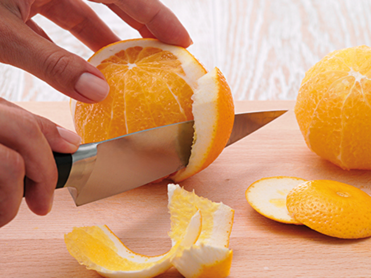 Eine Person schält mit einem Fissler-Küchenmesser eine Orange auf einer hölzernen Unterlage, wobei die geschälte Orange und die Schalen daneben liegen.
