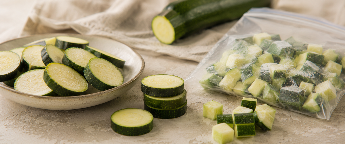 Frische Zucchini, in Scheiben geschnitten und auf einem Teller angerichtet, daneben ordentlich gestapelte Zucchini-Scheiben, im Hintergrund eine ganze Zucchini und fachmännisch gewürfelte Stücke in einem durchsichtigen Beutel – all dies unterstreicht die Präzision beim Schneiden mit den hochwertigen, in Deutschland entwickelten Küchenhelfern von Fissler.