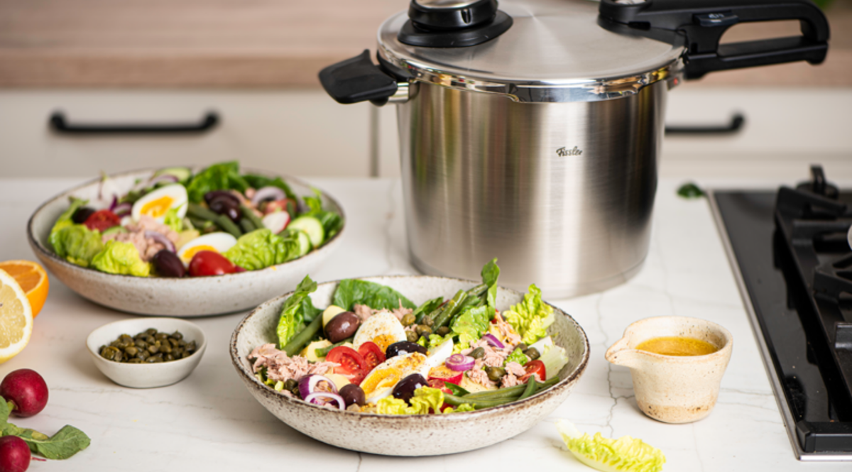 Ein Fissler Schnellkochtopf aus rostfreiem Stahl auf einer Küchentheke mit frischen Salaten, Kapern, Soßenkrügen und Gemüse.