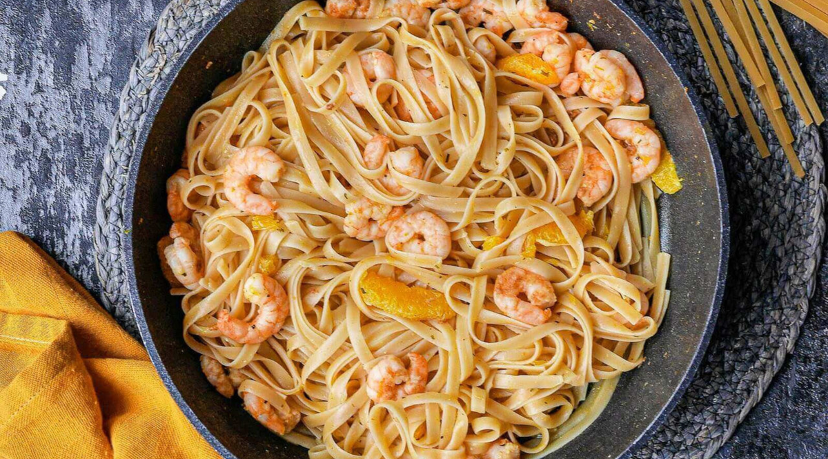 Eine hochwertige Fissler-Pfanne mit Linguine, Garnelen und Orangenscheiben auf einer strukturierten grauen Oberfläche, gelbe Serviette und Nudeln daneben.