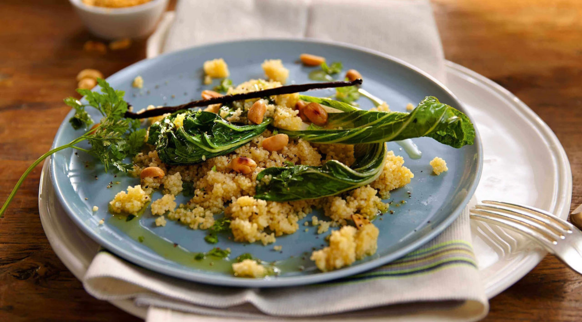 Couscous mit Grünzeug, Pinienkernen und Kräutern, serviert auf einem blauen Teller mit einer Fissler-Gabel aus rostfreiem Stahl auf einem Holztisch.