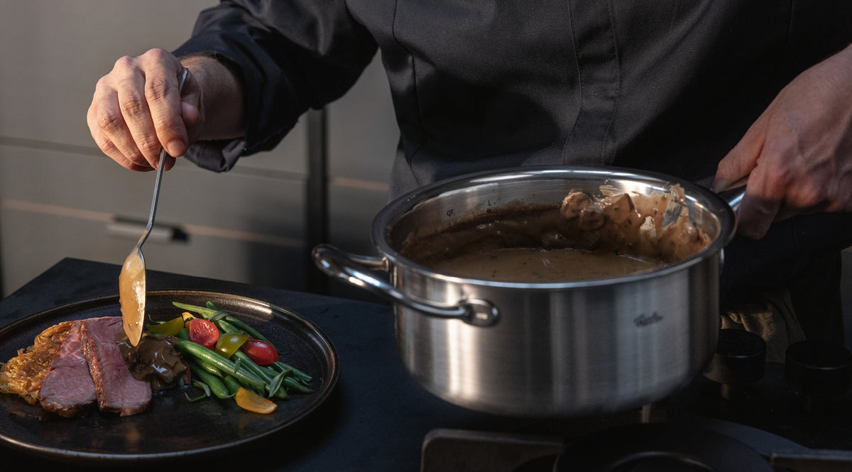 In einem Fissler-Edelstahltopf serviert man eine cremige Sauce über Fleisch, grüne Bohnen, Tomaten und Gemüse auf einem Teller.