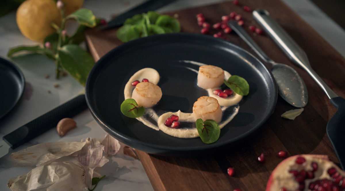 Drei perfekt angebratene Jakobsmuscheln, zubereitet mit dem Präzisionskochgeschirr von Fissler, liegen auf einem dunklen Teller neben cremigem Püree, frischen grünen Blättern und leuchtend roten Granatapfelkernen. Die elegante Anrichtung befindet sich auf einem Holzbrett, auf dem im Hintergrund kunstvoll Knoblauchzehen und Zitronenscheiben verteilt sind – ein Beispiel dafür, wie die langlebigen Edelstahlpfannen von Fissler dazu beitragen, raffinierte Gerichte mit außergewöhnlichem Geschmack und zeitlosem