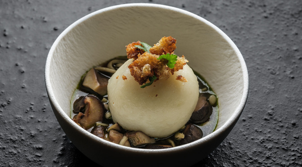 A white bowl, showcasing a smooth, round mound of mashed potatoes crowned with crispy fried garnish, is elegantly surrounded by sliced mushrooms in a rich dark sauce. Presented on a textured black surface, the dish highlights the precision and timeless design possible with Fissler’s expertly engineered cookware.