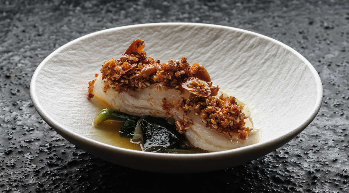 A perfectly cooked fillet of fish with a golden nut and herb crust rests on a bed of wilted greens, presented in a shallow, elegantly textured white bowl with a delicate sauce at the base—a result achieved with precision and care using Fissler’s expertly engineered cookware.