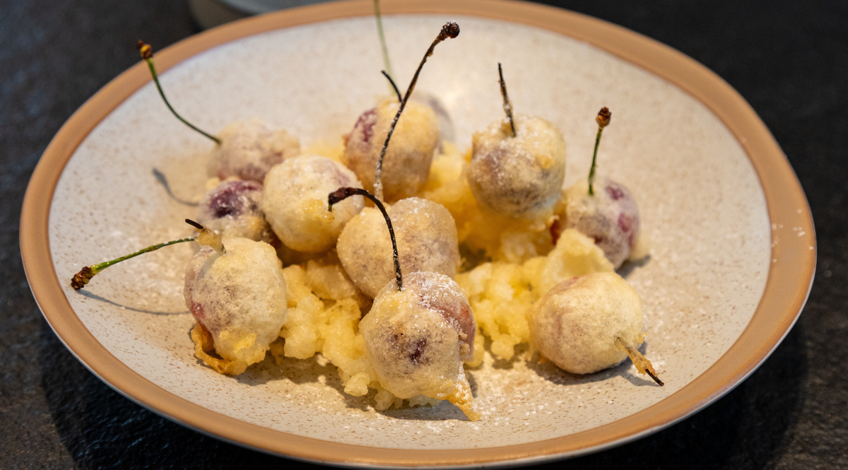 Cherries delicately coated in a light, crispy tempura batter, presented on a beige ceramic dish with a refined tan rim. Their vibrant stems remain attached, highlighting the elegance and precision that define the Fissler tradition of timeless German cookware craftsmanship.