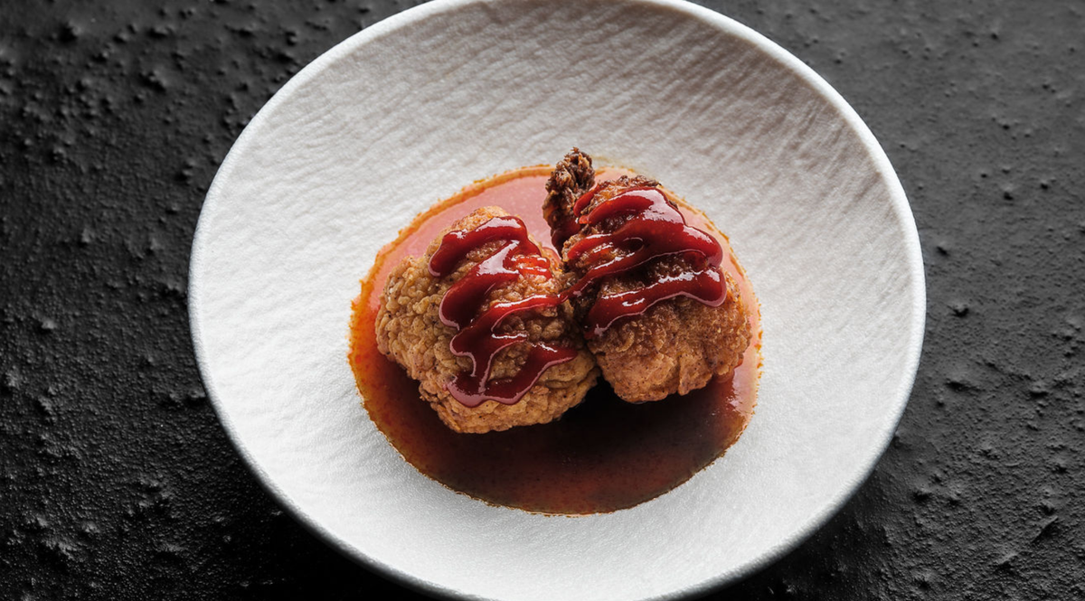 Two pieces of golden fried food, elegantly topped with vibrant red sauce, are artfully presented on a pristine white plate finished with a rich brownish sauce. The setting rests atop a textured black surface, highlighting the timeless design and premium quality associated with Fissler's German-engineered cookware.