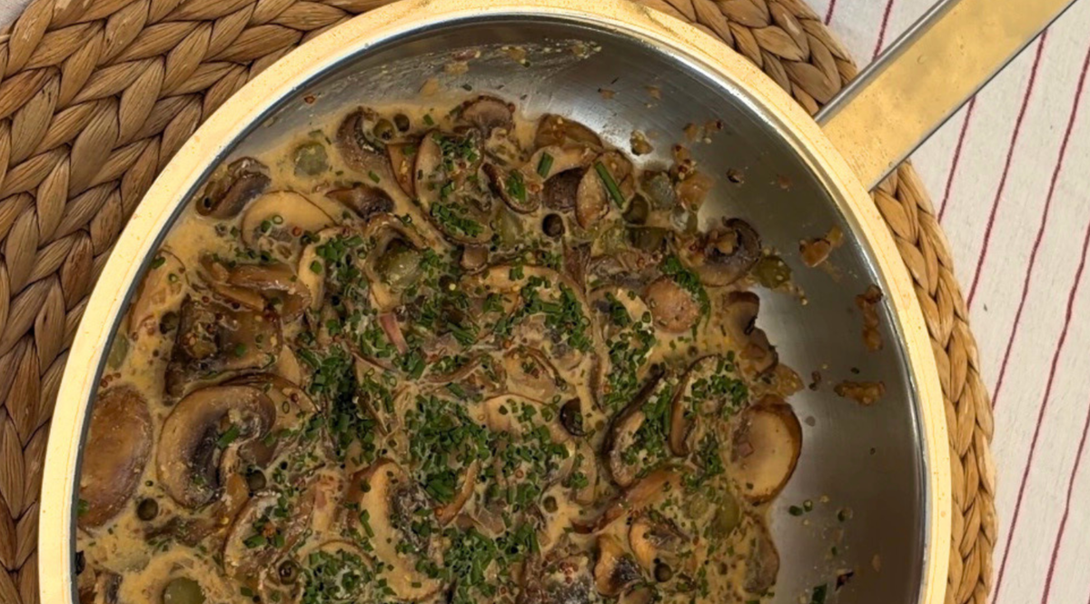 Cremig angebratene Pilze, garniert mit frisch gehackten Kräutern, serviert in einer hochwertigen Edelstahlpfanne von Fissler. Die Pfanne steht elegant auf einer gewebten Tischunterlage, unter der teilweise ein gestreiftes Tuch hervorblitzt – ein Detail, das die Präzisionstechnik und das zeitlose Design unterstreicht, die das in Deutschland hergestellte Kochgeschirr von Fissler auszeichnen.